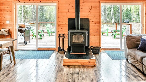 A spacious living room with a large black wood-burning stove, wooden floors, and large windows offering views of the outdoors.