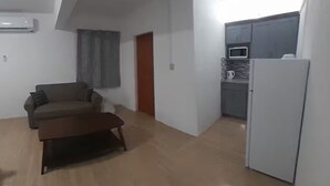 Un espacio habitable compacto con sofá, una mesita de centro, kitchenette y una puerta que da a otra habitación.