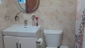 Un baño con lavamanos blanco, espejo y retrete. Hay artículos de tocador sobre el lavamanos, un cubo de basura y un pequeño bote de basura.