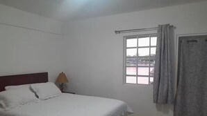 Un dormitorio con cama, mesita de noche, lámpara, ventana con cortinas y puerta.