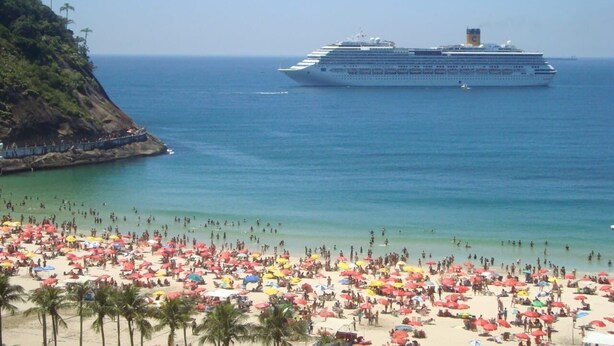 Un crucero amarrado cerca de una playa con numerosas sombrillas y gente.