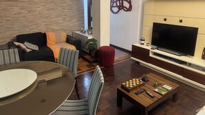 Una sala de estar moderna con un sofá, una mesa de centro con ajedrez y controles remotos, un televisor sobre un mueble bajo, y un aire acondicionado instalado en la pared.