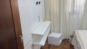Una habitación pequeña y limpia, con piso de madera, un lavamanos blanco, una mesita pequeña blanca y una cama con cubrecama blanco.