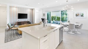 Eine moderne Küche mit Kochinsel, ein Essbereich mit rundem Tisch und Stühlen sowie ein Wohnbereich mit Sofa und Fernseher.
