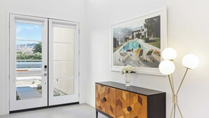 A room with a wooden cabinet, a framed picture on the wall, and a floor-to-ceiling sliding door with a view of a pool and mountains. - Lumen at Tahquitz: New Build With Pool, Fire Pit & Designer Interiors (Palm Springs)