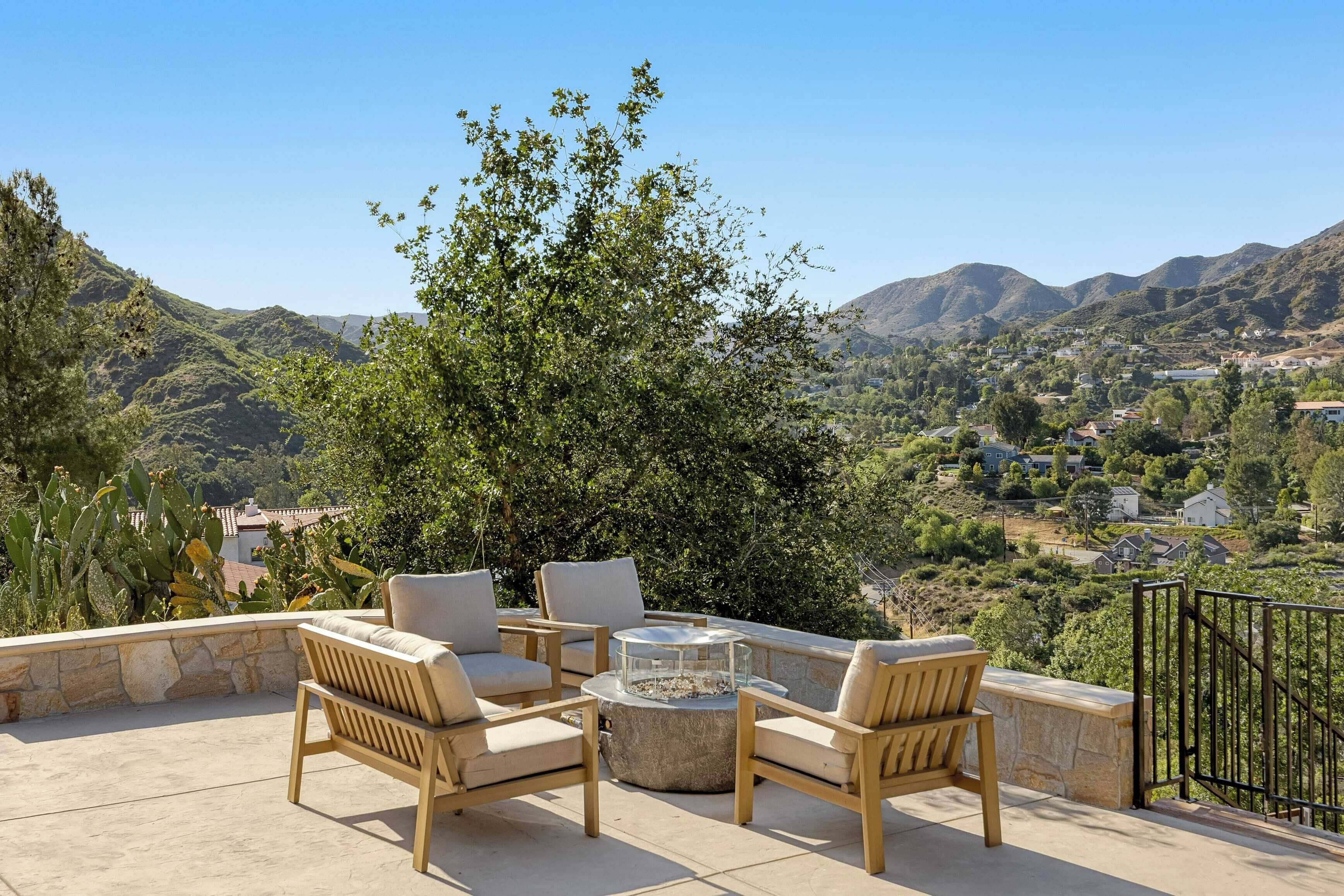 An outdoor patio with four wooden chairs and a glass-top table, overlooking a mountainous landscape with a view of a residential area.