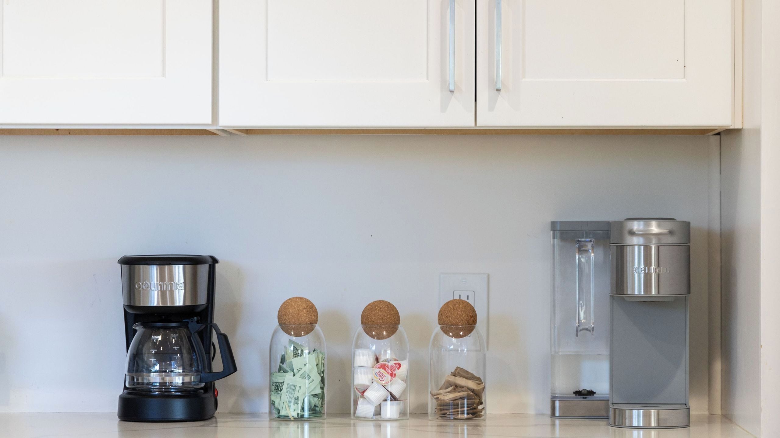 Una encimera de cocina con una cafetera, tres frascos de especias y un dispensador de agua.