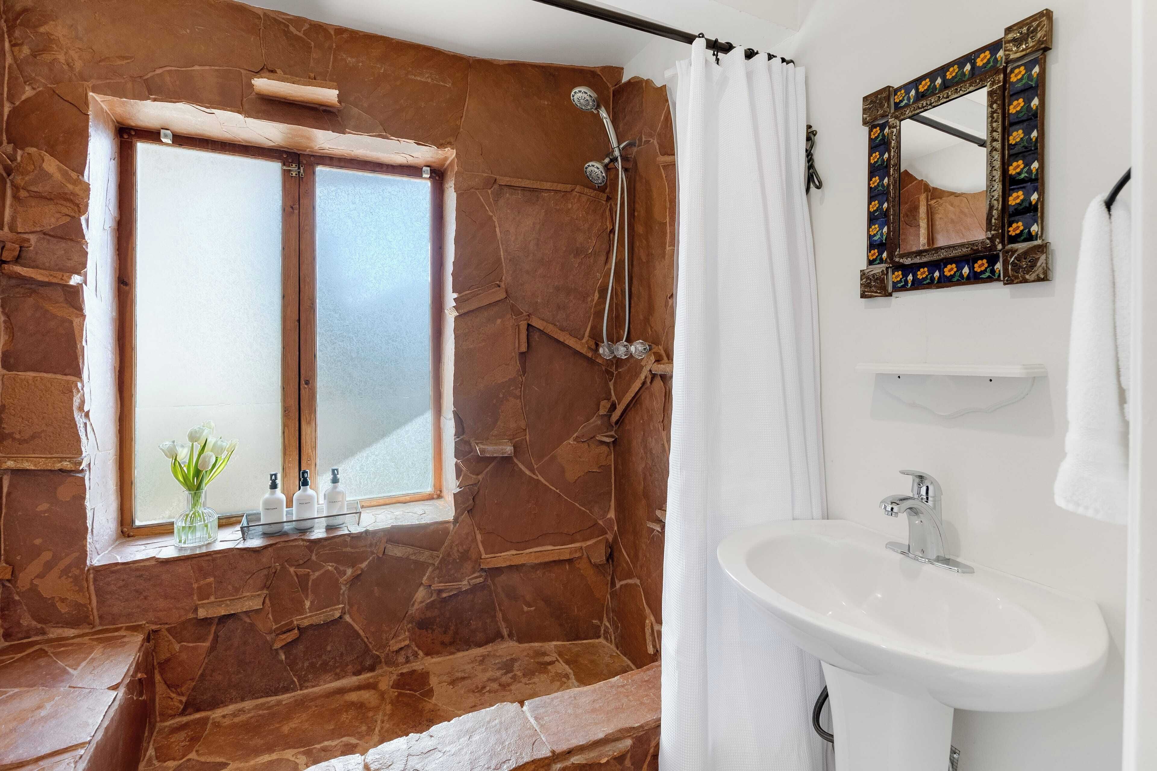 Un baño con pared de piedra, ventana con vistas, ducha, lavamanos y espejo.