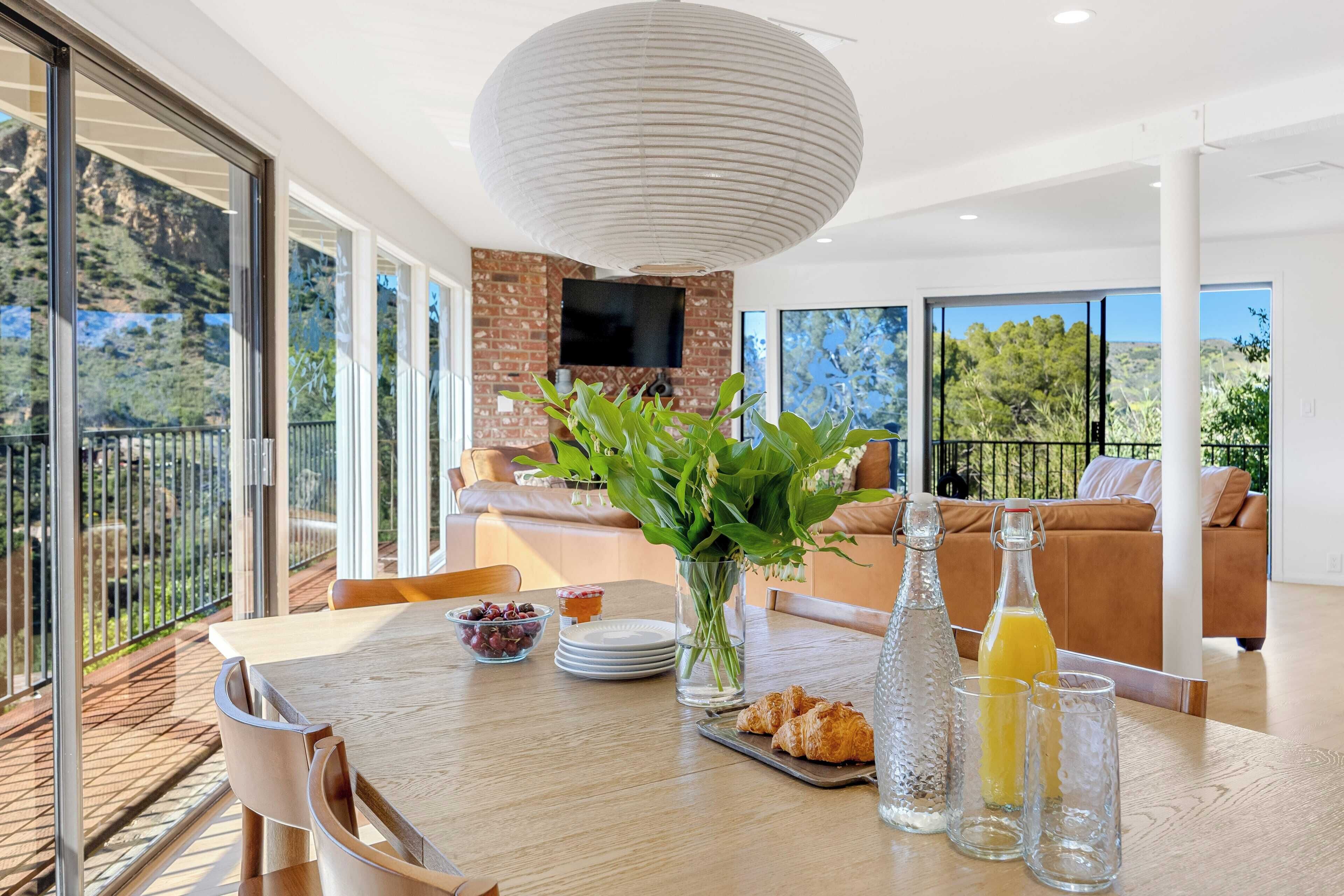 A modern dining area with a wooden table, clear glassware, and a vase of green plants.