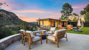 A patio with outdoor furniture and a fire pit, overlooking a mountainous landscape and a pool area.