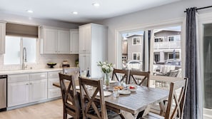 A modern kitchen with white cabinetry, a dining table with chairs, and a view of the neighborhood through large windows.