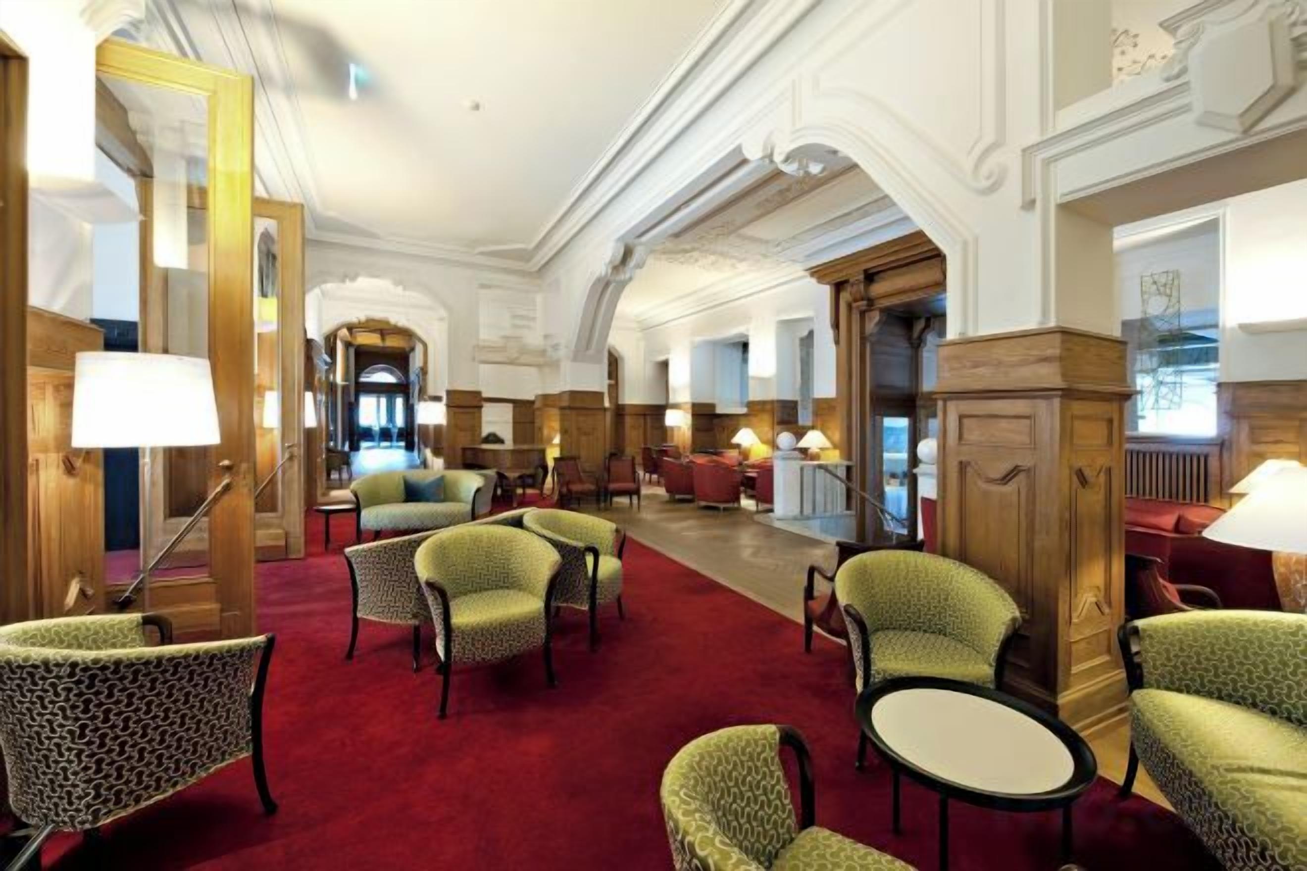 A hotel lobby with a red carpet, green upholstered chairs, and a grand staircase.