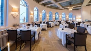 A spacious dining area with multiple tables and chairs, large windows, and a high ceiling with decorative elements.