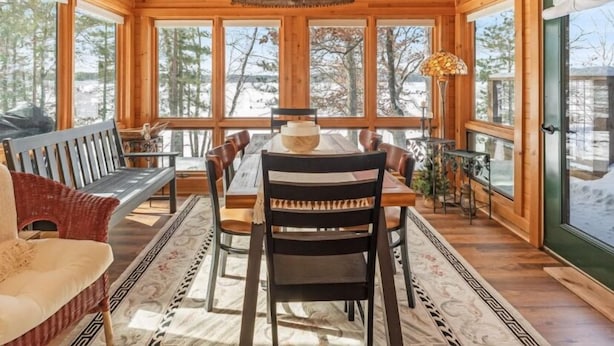 A cozy cabin interior with a wooden dining table, chairs, a bench, and a ceiling fan. Large windows offer views of the surrounding forest. - Charming 3-bedroom cabin with WiFi, AC in lovely Eagle River sleeps 8 people. (Eagle River)