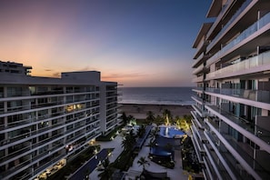 A modern hotel with multiple floors, a pool area, and a view of the ocean at sunset.
