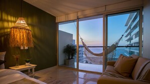 Une chambre d’hôtel moderne comprenant un lit, une table de chevet avec une lampe, un canapé, une grande porte-fenêtre coulissante donnant sur un balcon équipé d’un hamac et offrant une vue sur la mer.