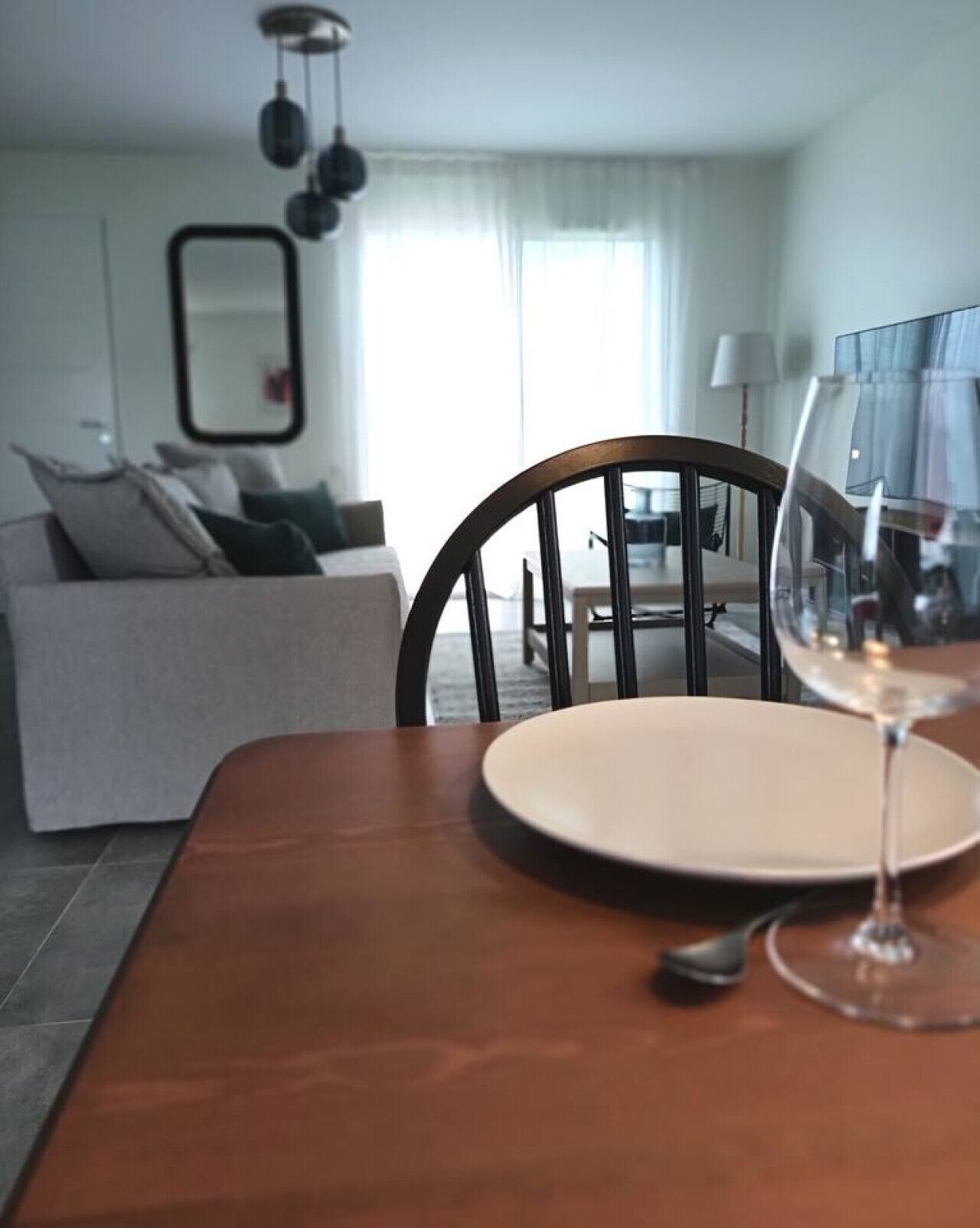 Ein Esstisch mit Weinglas und Teller, ein Holzstuhl und ein graues Sofa in einem modernen Wohnzimmer mit großem Fenster und einer Pendelleuchte.