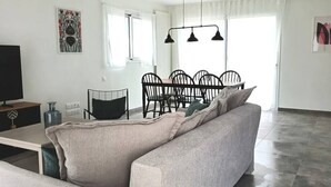 Un salon moderne avec un canapé, une table à manger et des chaises. Des lampes suspendues sont placées au-dessus de la table, et un téléviseur à écran plat est posé sur un meuble.