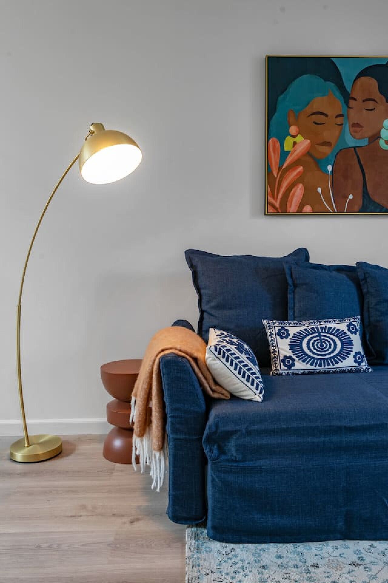 A room with a blue sofa, a patterned throw blanket, and a decorative pillow. A floor lamp with a gold base is positioned to the left.