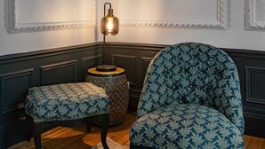 Une pièce dotée d’un parquet en bois foncé, d’un fauteuil à motifs, d’un pouf assorti, d’une petite table ronde avec une lampe et d’une œuvre d’art encadrée accrochée au mur.