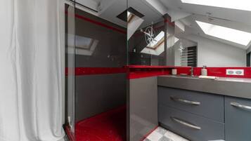 Un baño moderno con cabina de ducha de vidrio, una pared de acento roja y un tragaluz sobre la zona del lavamanos.