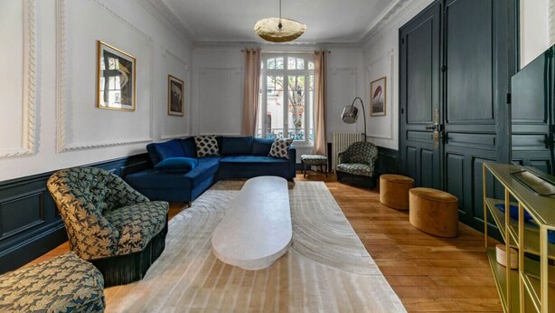 Un salon spacieux avec un grand canapé bleu, des fauteuils à motifs, un pouf blanc et un plancher en bois.