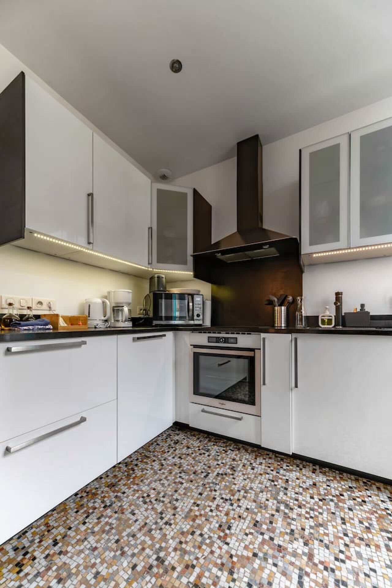 A modern kitchen with white cabinets, a black range hood, a microwave, and an oven. The floor has a mosaic tile pattern.