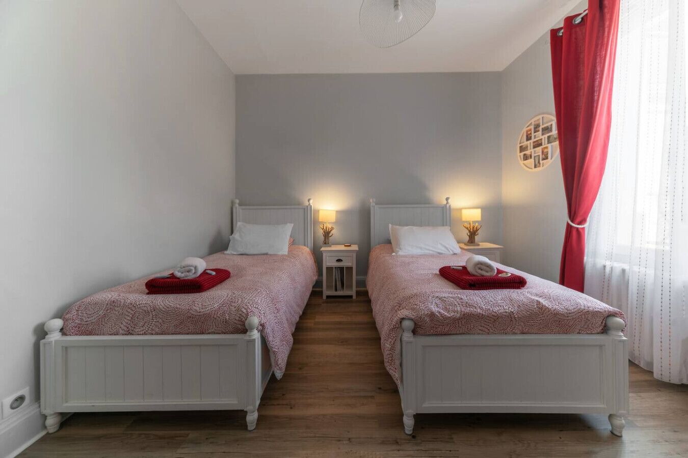 Quarto duplo com duas camas, cada uma com cabeceira branca, manta vermelha e toalha. Há mesas de cabeceira com luminárias, cortina vermelha e um quadro pendurado na parede.