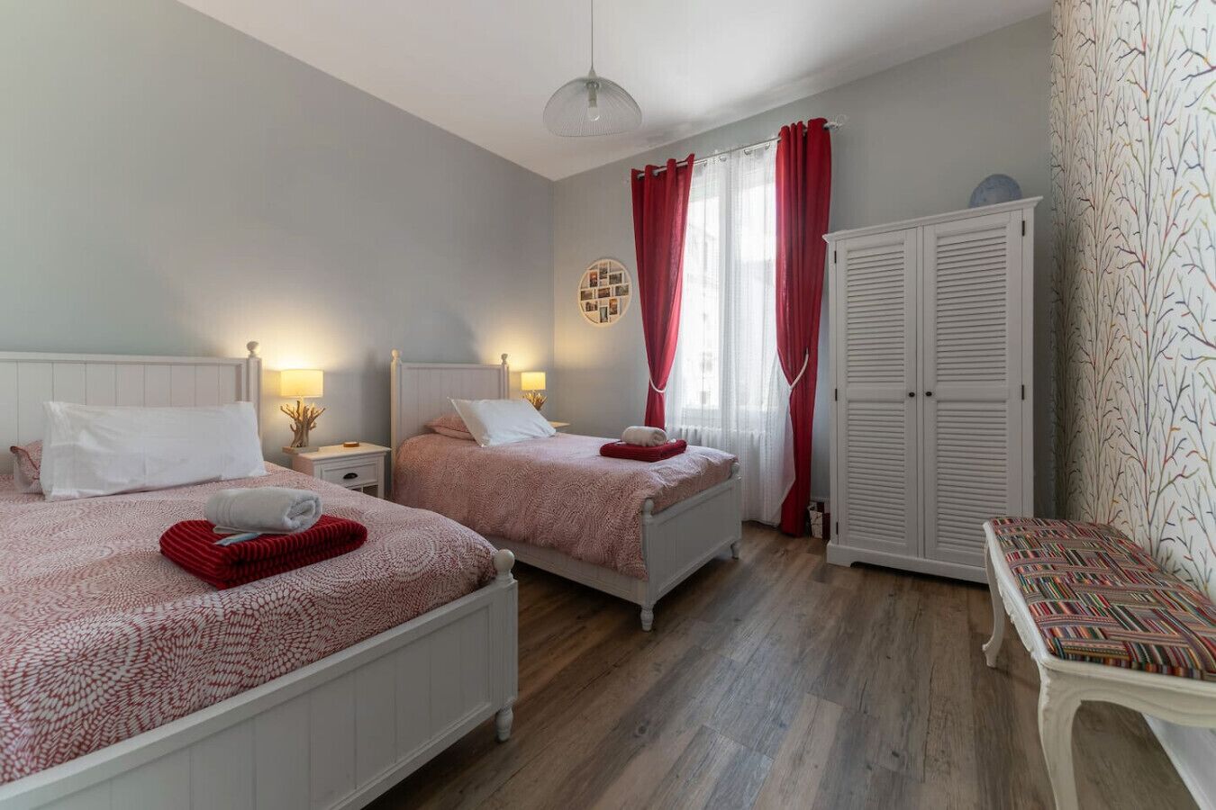 Une chambre avec deux lits, une armoire, une table de chevet avec une lampe et une fenêtre avec des rideaux rouges.