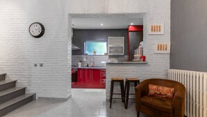 Una cocina moderna con armarios rojos, una pared de ladrillo blanco y un reloj. Hay dos taburetes y una silla marrón en la sala de estar.