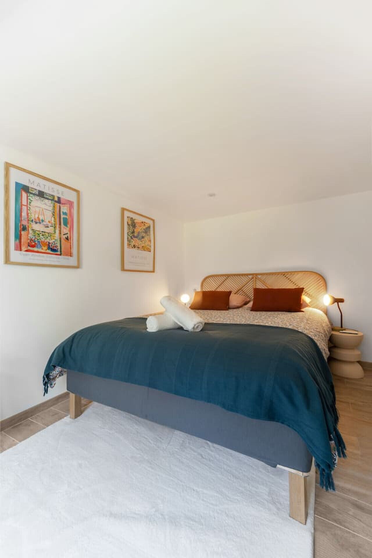 A bedroom with a bed, pillows, a bedside lamp, and two framed artworks on the wall.