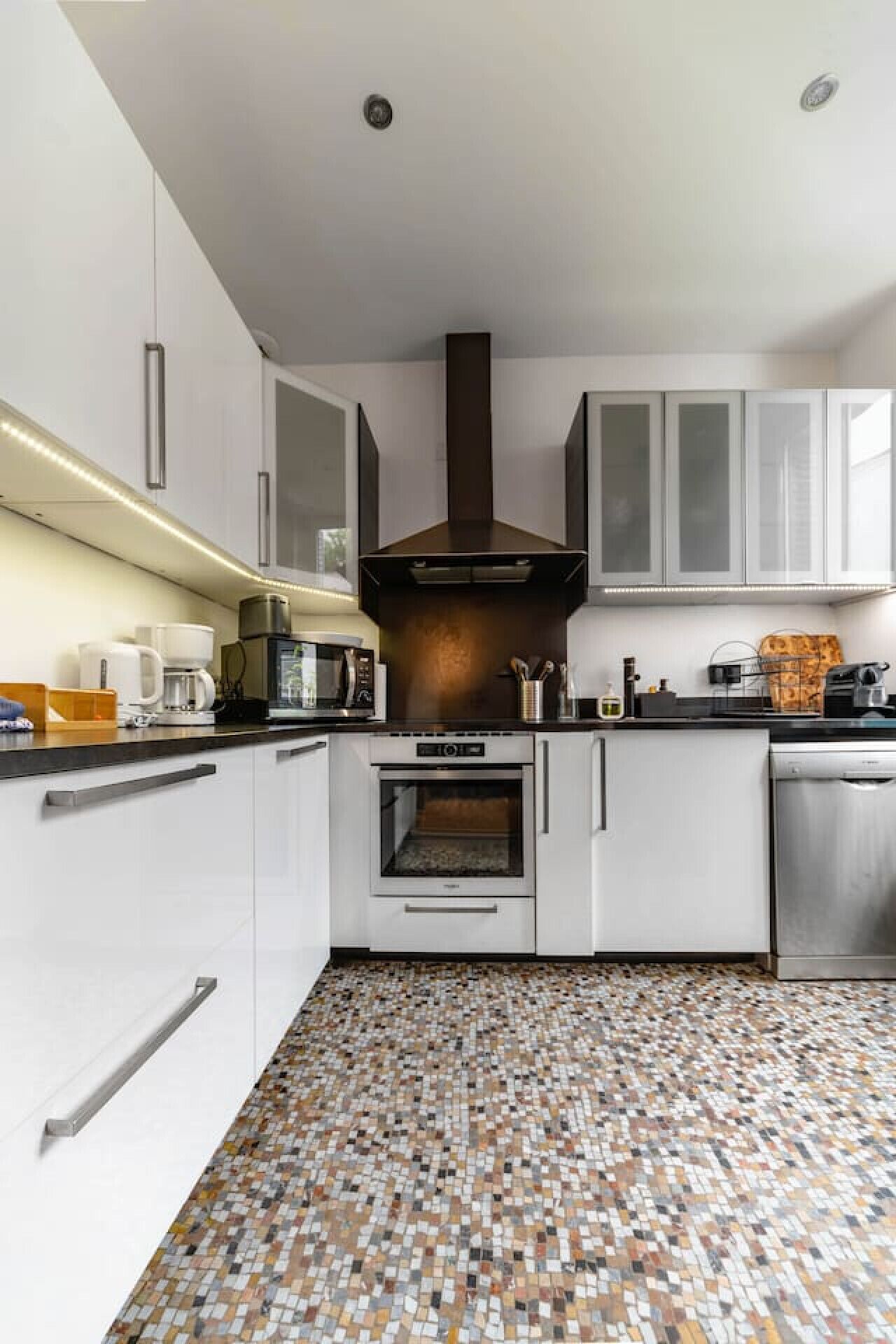 Une cuisine moderne avec des armoires blanches, une hotte de cuisine centrale, un four encastré et un sol carrelé à motifs.