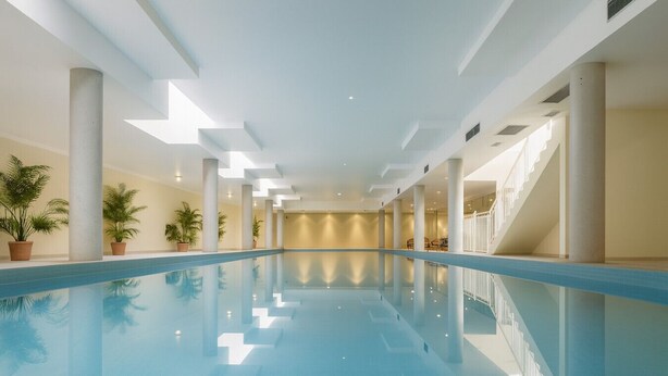 Una piscina interior larga con agua cristalina, rodeada de columnas blancas y plantas en macetas.