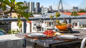 Mesa de comedor en la azotea con platos, vasos y un cuenco de uvas, con vistas a un paisaje urbano con edificios modernos y un puente.