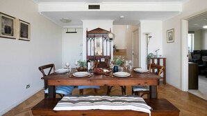 Un comedor con una mesa de madera dispuesta para seis personas, flanqueada por sillas. Un banco se ubica al pie de la mesa. Las paredes son blancas y el suelo es de madera.