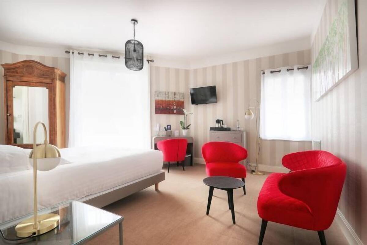 A hotel room with a bed, a desk, two red chairs, a TV, and a large mirror.
