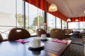 Een restaurantgedeelte in een hotel, met tafels voor twee personen, stoelen en een uitzicht op de stad door grote ramen.