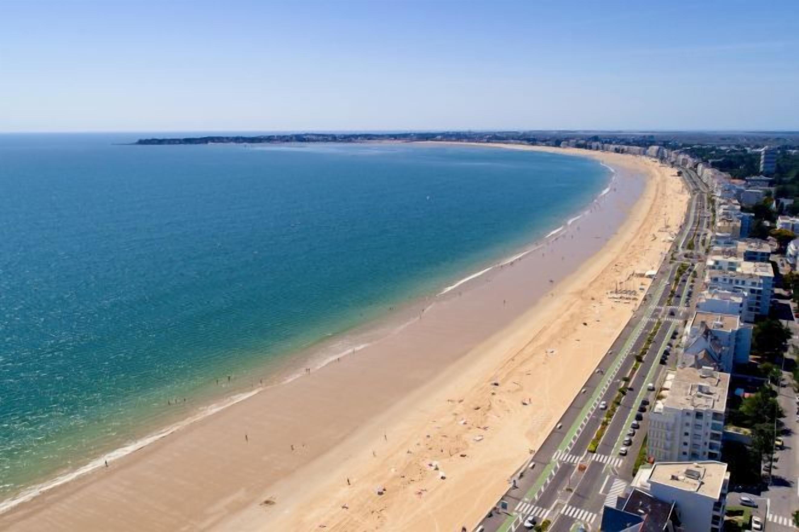 A coastal cityscape with a wide sandy beach, a clear blue sea, and a row of buildings along the shore.
