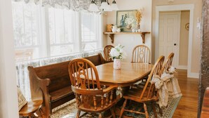 Une salle à manger avec une table et des chaises en bois, un banc, des rideaux en dentelle, un lustre et un tableau au mur.