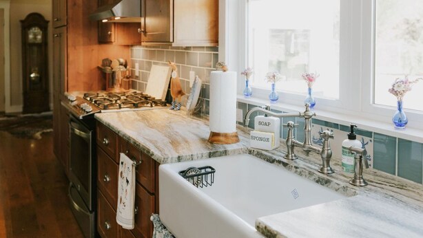 Une cuisine avec des meubles de rangement en bois, une cuisinière, un évier doté de distributeurs de savon et d’éponges, et une fenêtre ornée de fleurs.