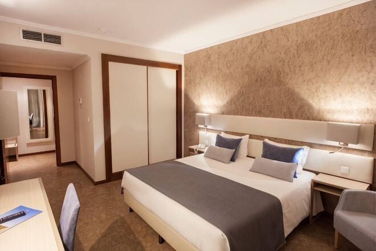 A hotel room with a bed, bedside tables, a desk, a chair, and a wardrobe.