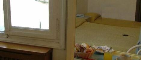 A hotel room with a bed, a window, a mirror reflecting the room, and a table with a bowl of fruit.