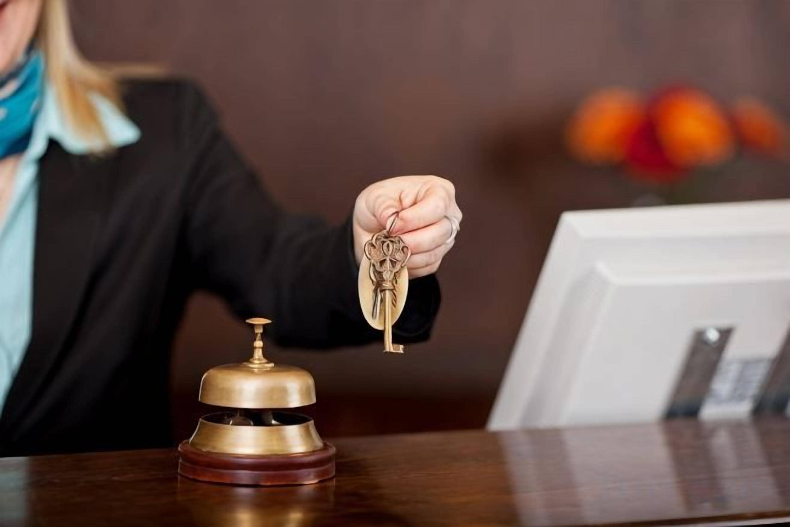 Eine Person hält einen Schlüssel vor einem Hotelrezeptionstresen mit einer Glocke und einem gerahmten Bild.