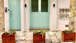 Un edificio con una puerta azul claro y una ventana, macetas de madera con plantas en la parte delantera, y cajas eléctricas en la pared.
