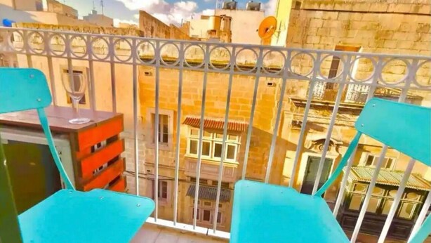 Un balcón con sillas turquesas y una pequeña mesa, con vistas a un paisaje urbano de edificios tradicionales.