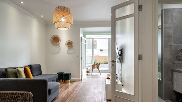 Un salon moderne avec un canapé gris, deux poufs verts, un fauteuil en osier et un luminaire suspendu.