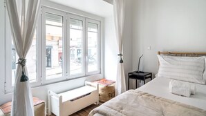 Une chambre à coucher avec un lit, une table de chevet, une fenêtre avec des rideaux, un radiateur et un petit banc.