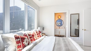A modern bedroom with a large bed, a view of the city, a bathroom with a mirror, and a round wall decoration.