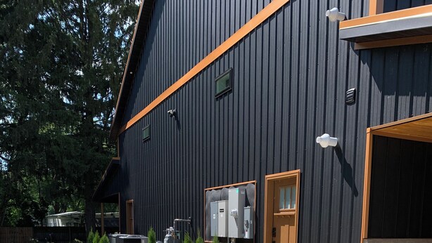 A modern building with a dark exterior, a wooden door, and a garage. There are outdoor electrical boxes and a pathway leading to the entrance. - Charming and Cozy Retreat in Newaygo, MI- The Rustic Barn dominium (Newaygo)