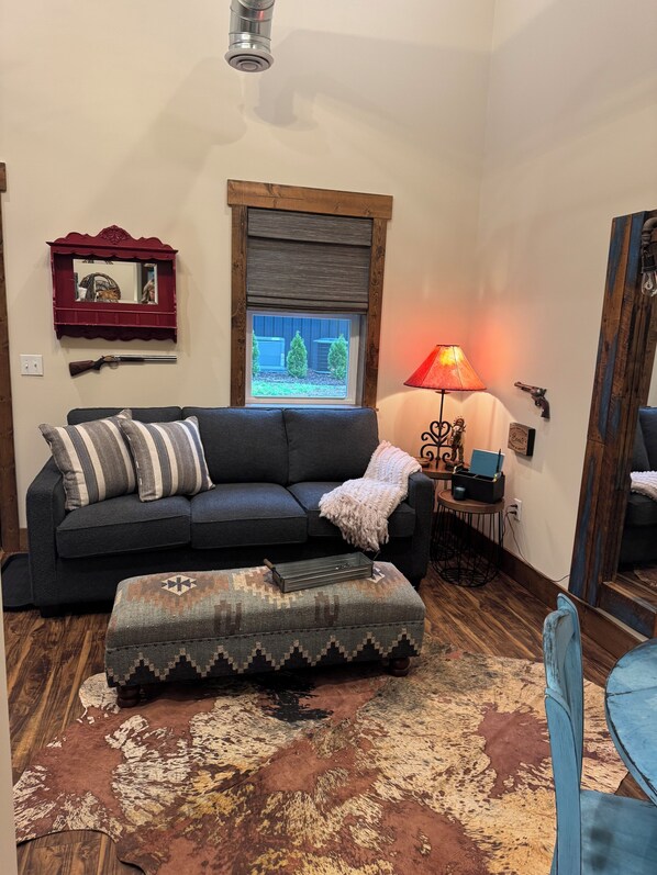 A living room with a dark grey sofa, striped pillows, a patterned ottoman, a red-framed mirror, a window with a view of trees, a red lamp, and a small table with a pistol and a book. - Charming and Cozy Retreat in Newaygo, MI- The Rustic Barn dominium (Newaygo)